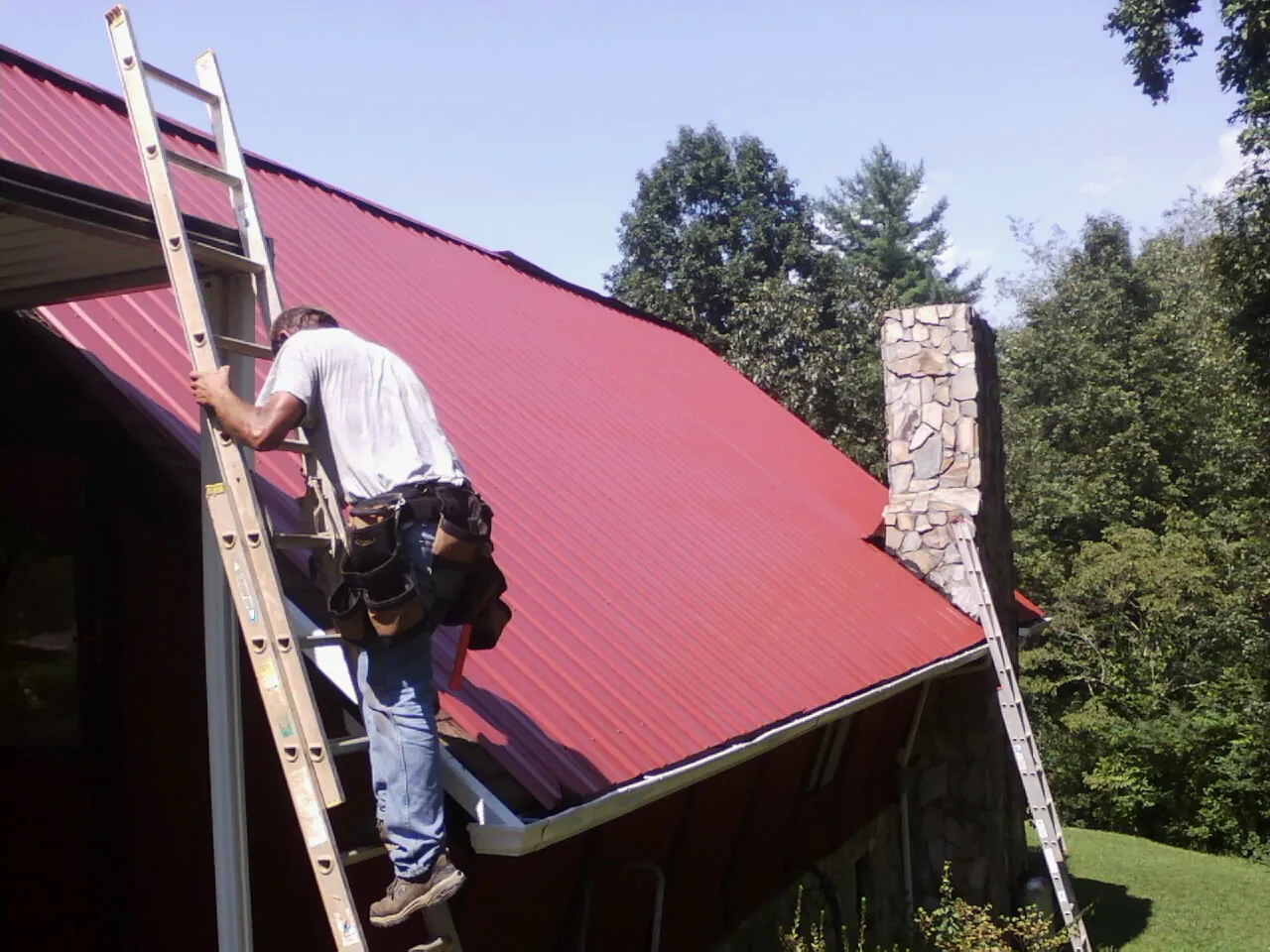 Roofer on a ladder beside a red metal roof and stone chimney — Soffit & Fascia Repair by Roofing Brothers in Rodessa